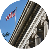 American flag flying above a neoclassical building with ornate columns against a clear blue sky.